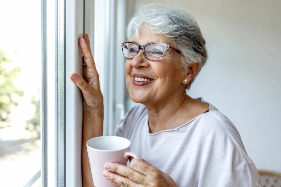 An older woman smiling while holding a mug by a window